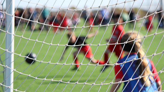 photo taken behind the goalie net, a young boy in black shirt and red shorts about to kick a soccer ball at the young girl goalie, caution to protect youth sports league from fraud