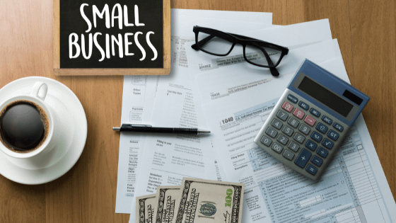 A calculator, pair of reading glasses, money and a chalkboard reading "Small Business," sitting on top of financial documents; image used for blog post about tax breaks for small businesses