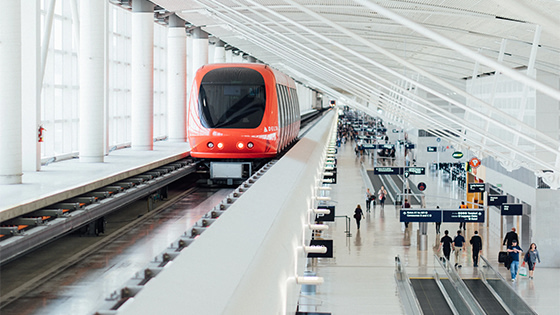 a red monorail train speeding overhead people walking through a transportation terminal; image used for UBIT transportation fringe benefits blog post