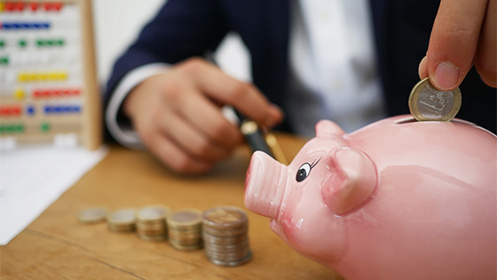 man in a business suit putting a silver coin into a pink porcelain piggy bank, next to stacks of coins, avoiding excess benefit transactions to keep tax-exempt status
