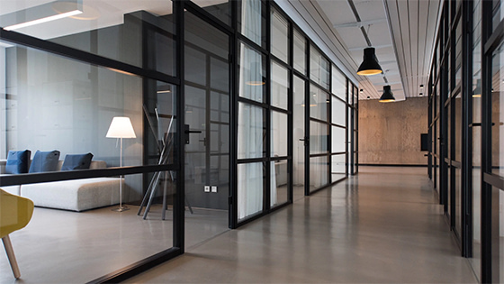 office hallway with black-frame glass doors and windows, in one room sits a white couch with three navy blue pillows, overhead lamps hang from the ceiling, possible leasing option for not-for-profit organizations