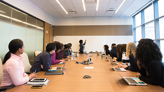 a group of people sit in a conference room around a wooden desk with someone leading discussion at the table head writing on a whiteboard; image used for blog post about in-person not-for-profit board retreats