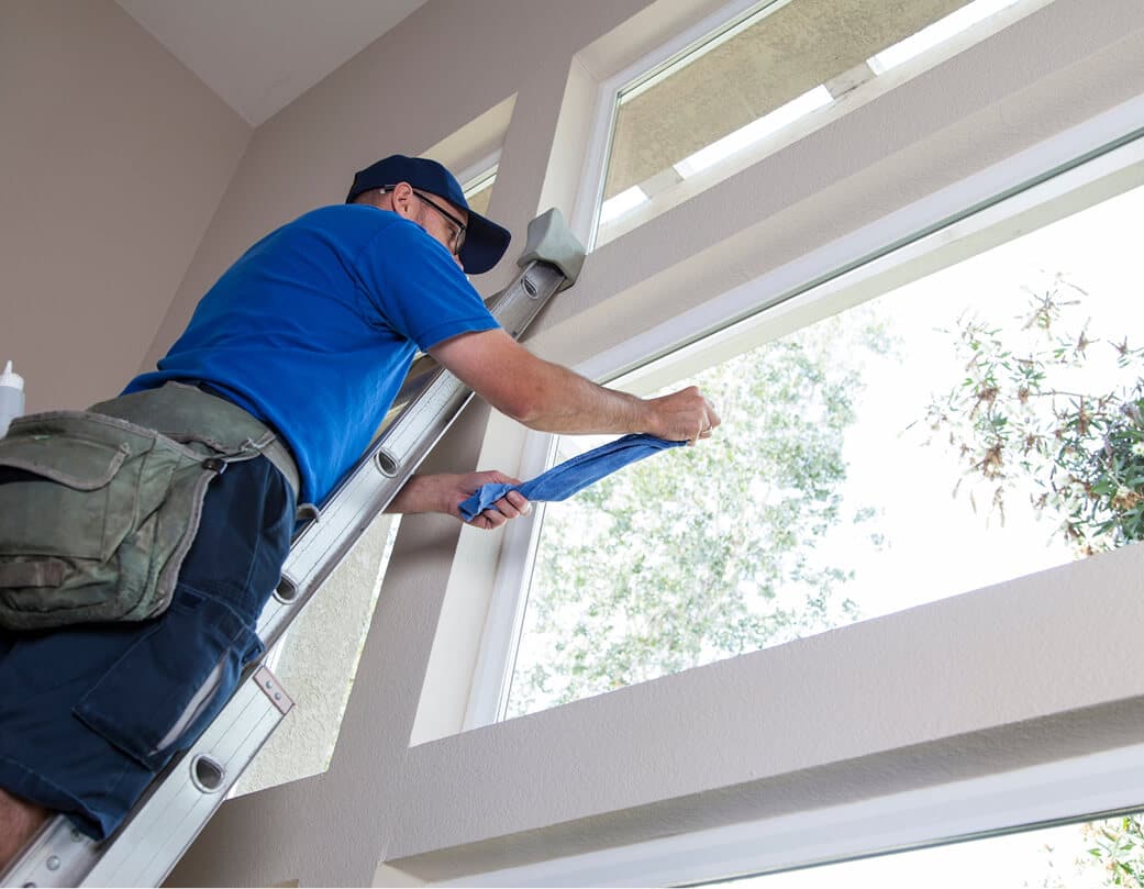 Professional window cleaner wiping tall living room windows from a ladder
