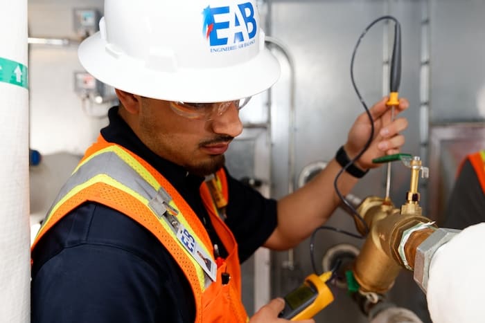EAB technician wearing a hard hat and safety vest, using a digital manometer to measure airflow in an HVAC system