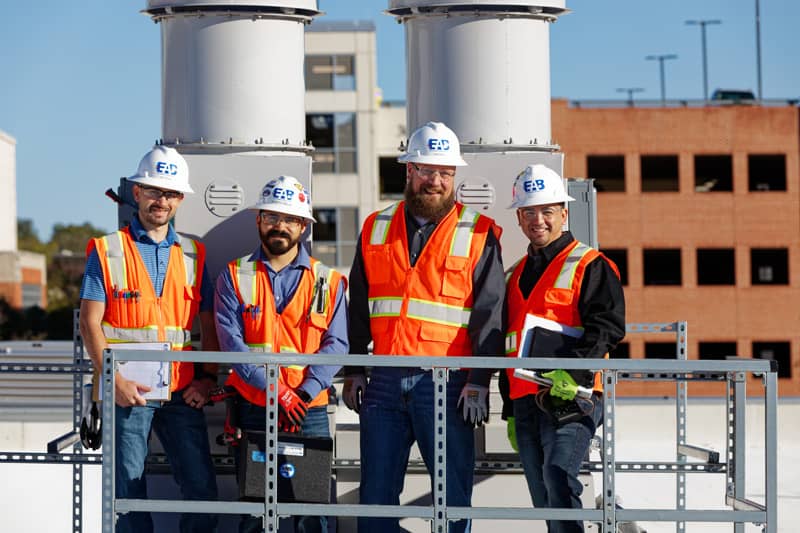 Four commissioning engineers from EAB pose on a rooftop, standing in front of industrial air vents, with a clear blue sky and a building in the background
