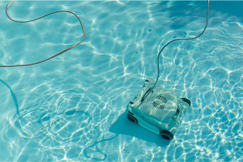 A white robotic pool vacuum is shown on the bottom of a swimming pool, with its cord leading to the surface