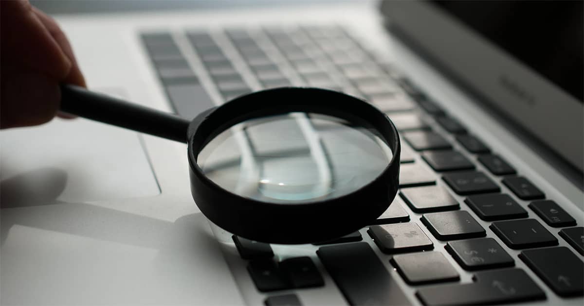 someone holding a magnifying glass to a laptop keyboard; image used for blog post about Danielle Supkis Cheek article about fraud prevention