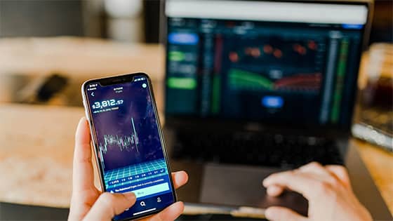 a person holds a cell phone with the screen showing a graph and "$3,812.57" in their left hand and in the background is a laptop with another graph with green and red lines; portrayal of selling securities and avoiding wash sale rule
