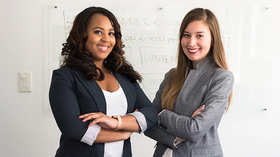 a Black woman and American woman pose together, wearing blazers, in front of a white board; image used for blog post about not-for-profits classifying workers for tax purposes
