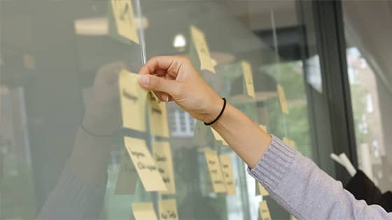 woman in long-sleeve grey shirt putting a yellow post-it note on a dry erase board; image used for blog post about not-for-profit leaders delegating tasks