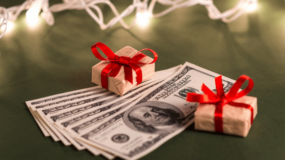 An image of one-hundred dollar bills and gift-wrapped boxes sitting in front of holiday lights