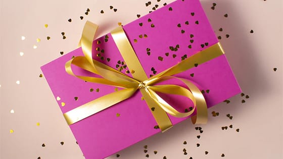 a box wrapped in pink paper with a gold ribbon tied into a bow, sprinkled with heart-shaped gold confetti; image used for blog post about proposed amendments for NFP nonfinancial assets from FASB