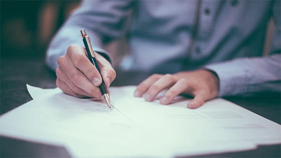 man in a blue button down shirt holds a pen to paper to sign documents; image used for a blog post about the CARES Act affecting retirement plans and charitable donation rules