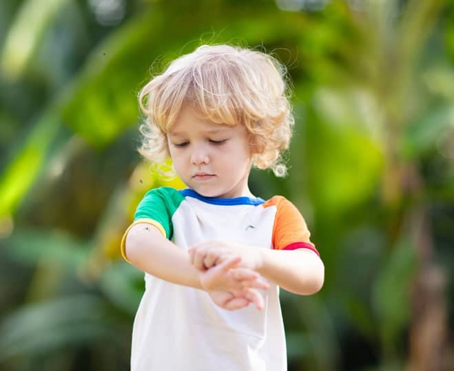 A young child with curly blonde hair wearing a colorful shirt, looking down at a mosquito on their arm in a lush green outdoor setting