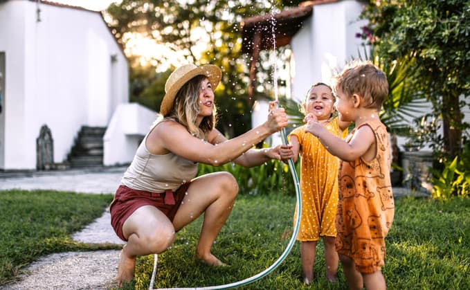 A woman and two young children playing with a garden hose in a backyard, enjoying the water on a sunny day