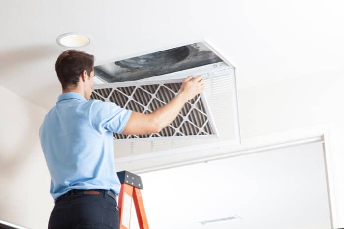 a hvac professional standing on a ladder, removing an air filter from a ceiling air duct