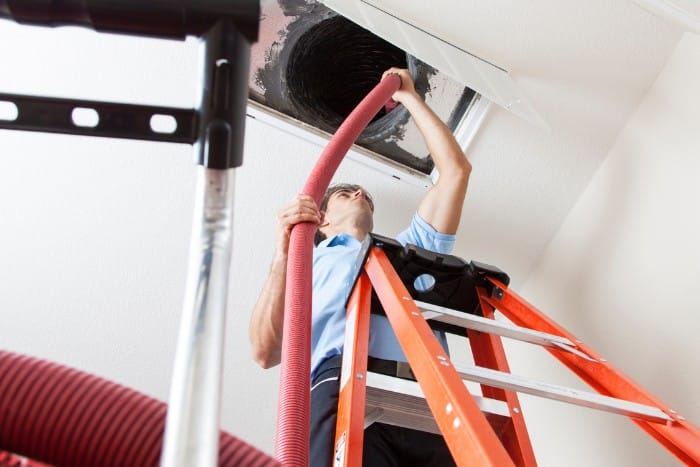 A technician using specialized equipment to clean air ducts while standing on a ladder