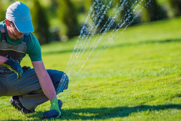 A professional adjusting a lawn sprinkler on a green lawn, with water spraying in multiple directions